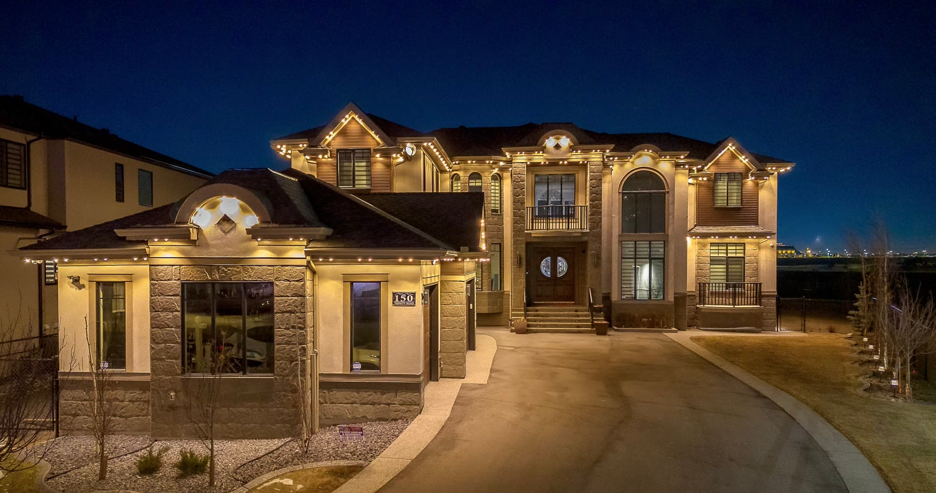 Large estate home traced in warm-white permanent lights at dusk, rooflines and gables evenly lit